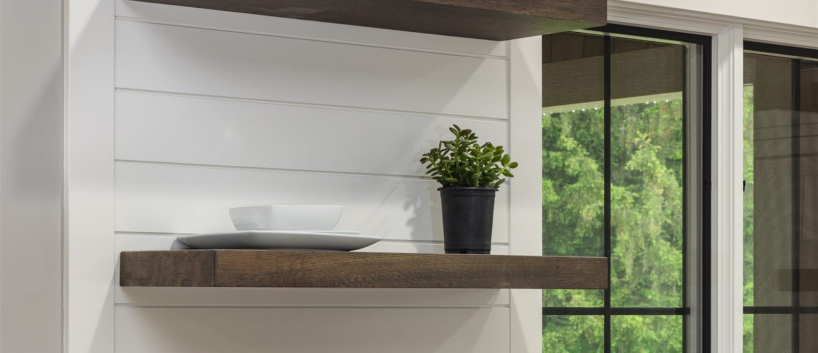Dark-stained floating shelf against a plain shiplap wall, styled with plates and a succulent, with a window view