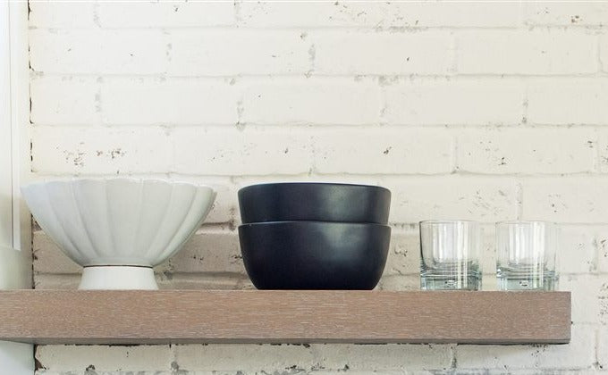 Floating shelf with a white ceramic bowl, black bowls, and drinking glasses against a whitewashed brick backsplash