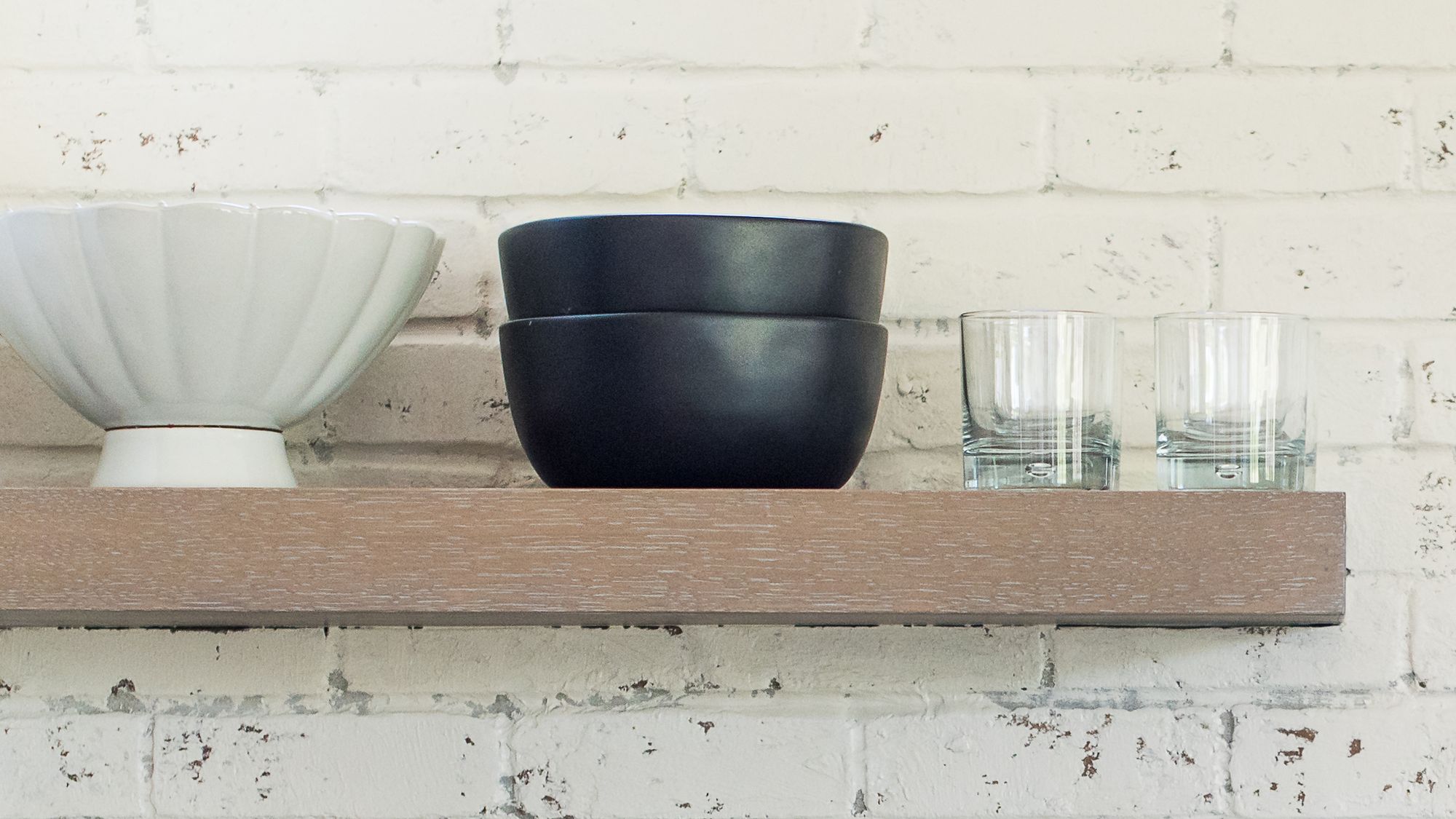 Light oak floating shelf holding a white ceramic bowl, two black bowls and two glasses on a whitewashed brick kitchen wall